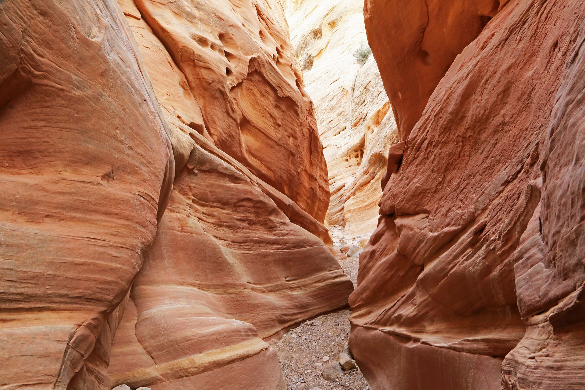 A narrow slot canyon featuring towering, smooth, orange-red sandstone rock walls and a rocky, gravel-covered floor.