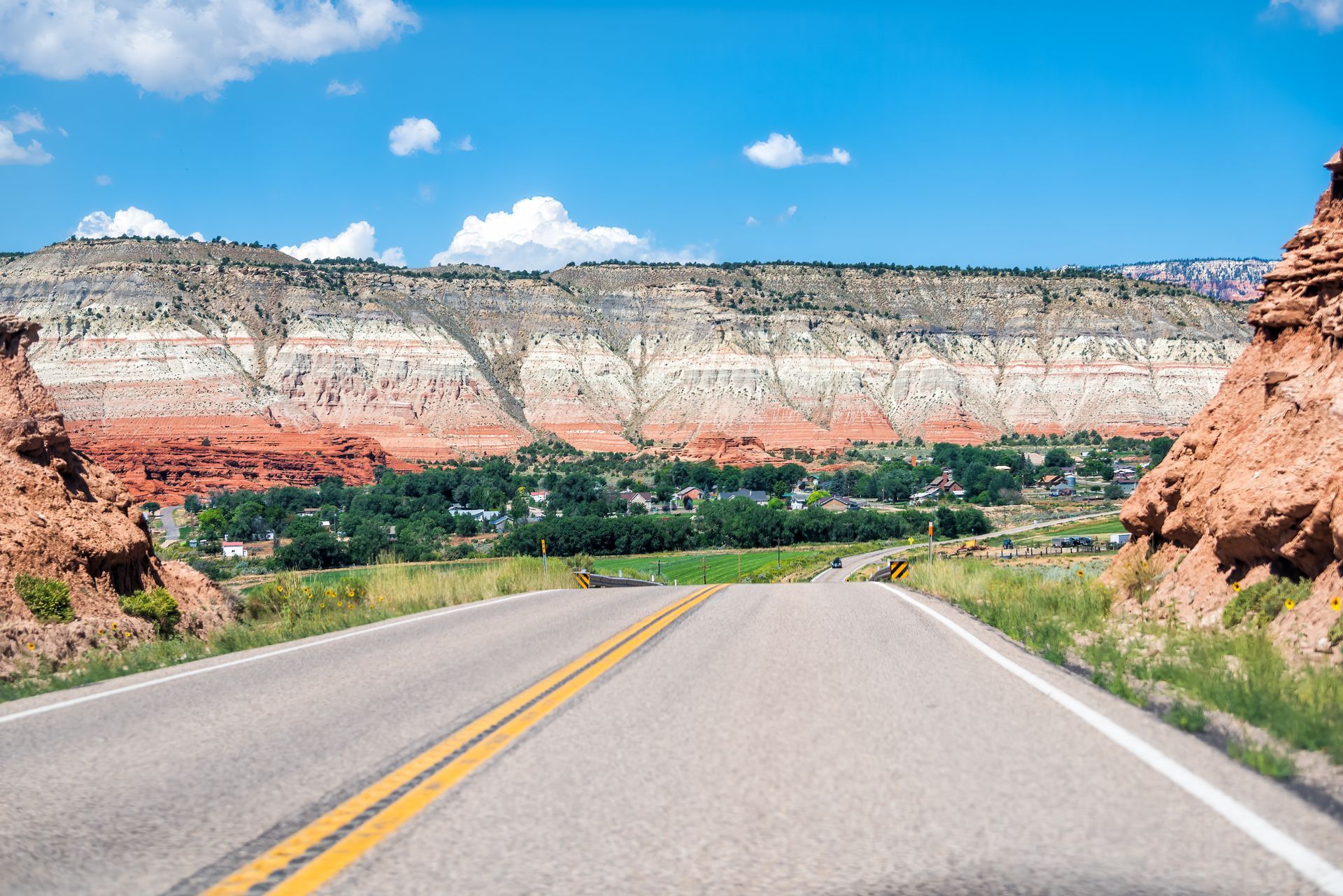 A paved road leads toward a green valley nestled beneath a long, multi-layered cliff face under a clear blue sky.