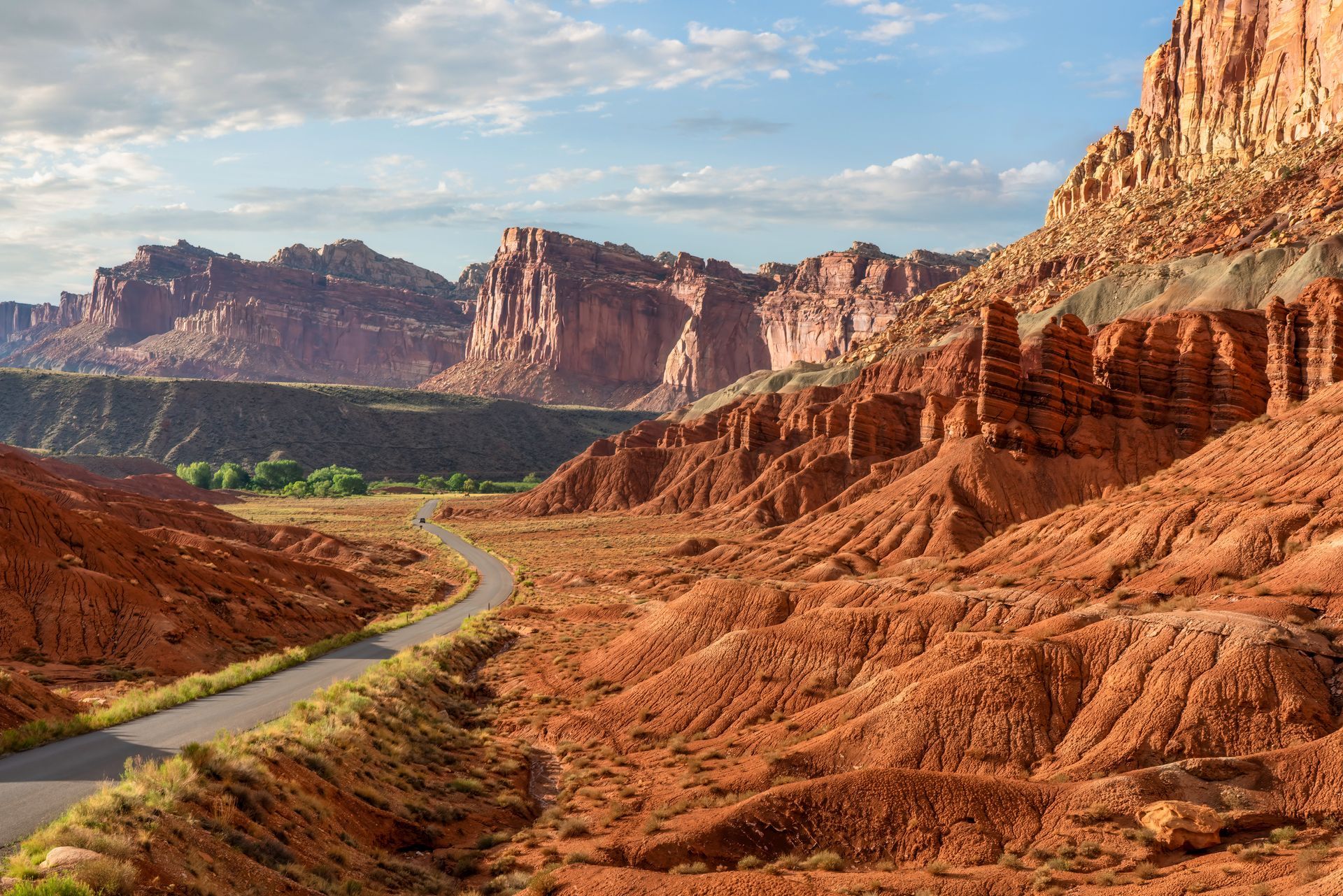 A winding road cuts through a desert landscape with reddish rock formations and cliffs under a blue, cloudy sky.