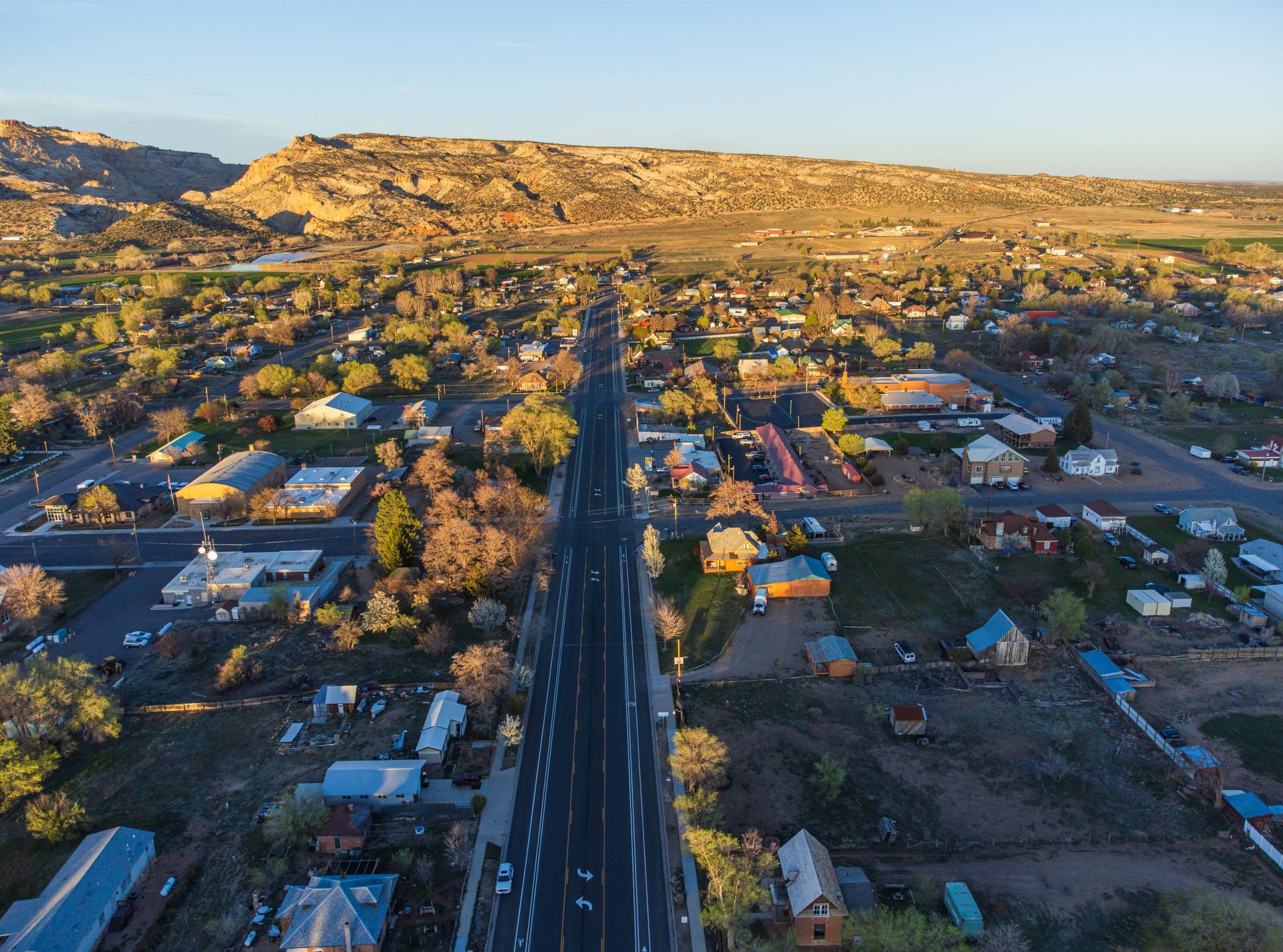 An aerial view shows a town road stretching toward a sunset-lit canyon backdrop, surrounded by houses and trees.