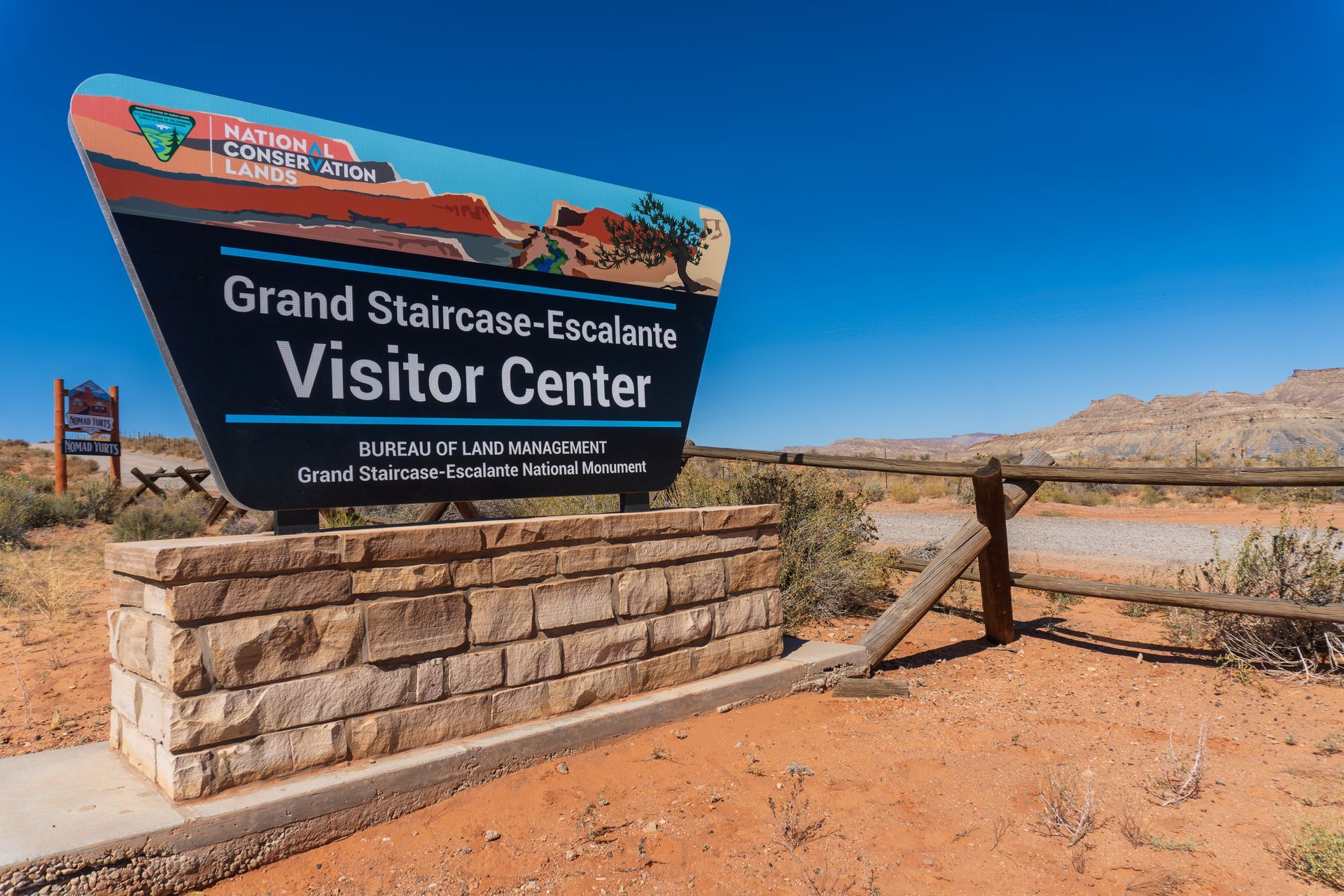 Grand Staircase-Escalante Visitor Center sign on a stone base, set in a desert landscape under a clear blue sky.