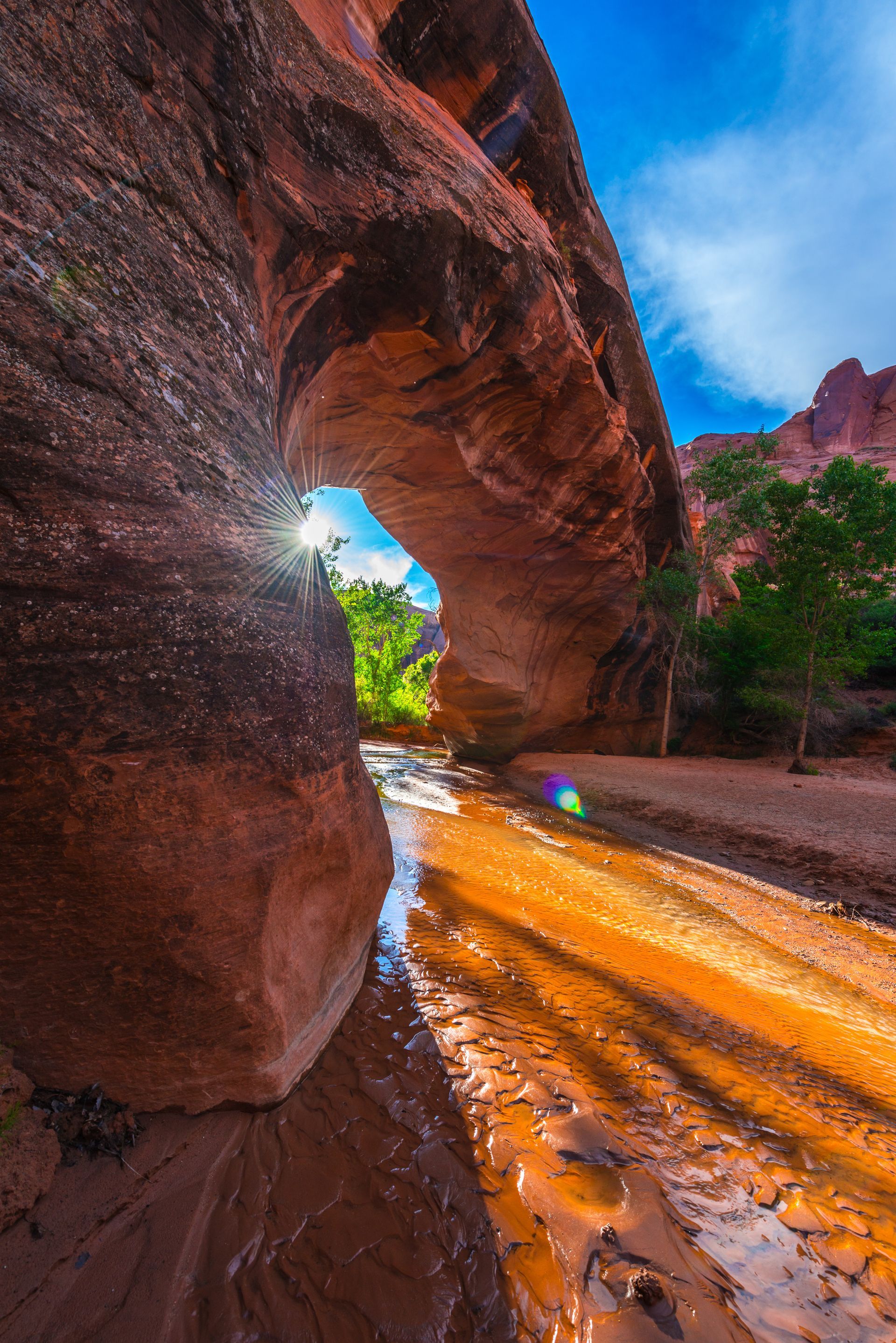 A natural sandstone arch frames a sunlit canyon stream with vibrant orange, red, and golden hues under a blue sky.