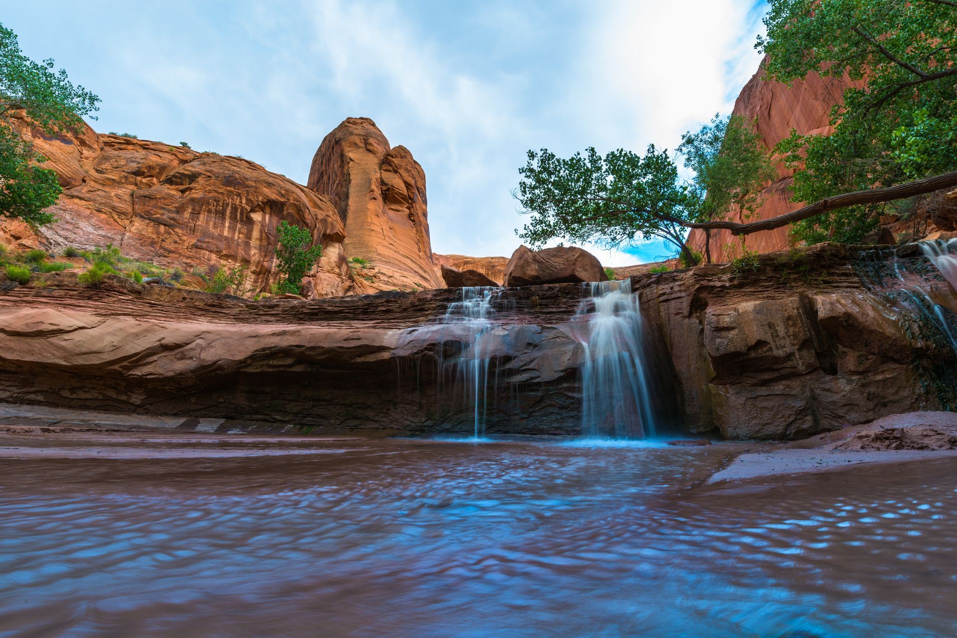 A small, gentle waterfall cascades over red rock ledges into a stream, set against a backdrop of desert cliffs and trees.