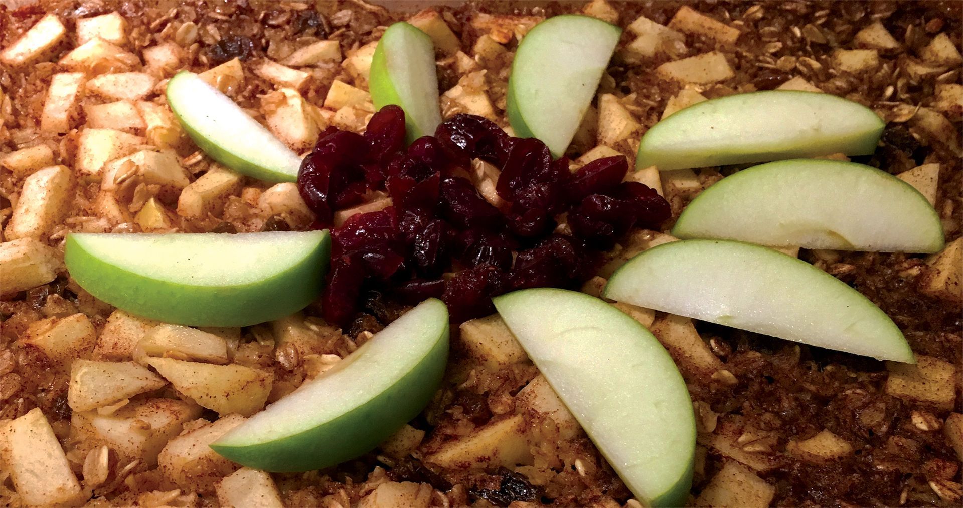 A baked apple crisp dish topped with a garnish of fresh green apple slices arranged in a circle around dried cranberries.