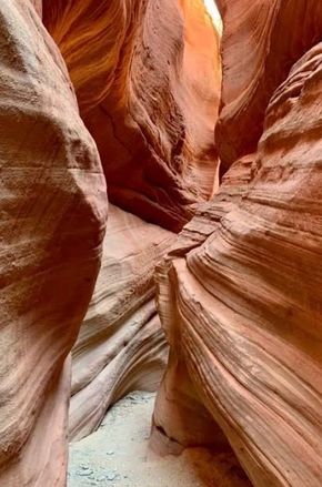 A narrow, winding slot canyon with smooth, wavy orange sandstone walls and a sandy floor under soft overhead light.