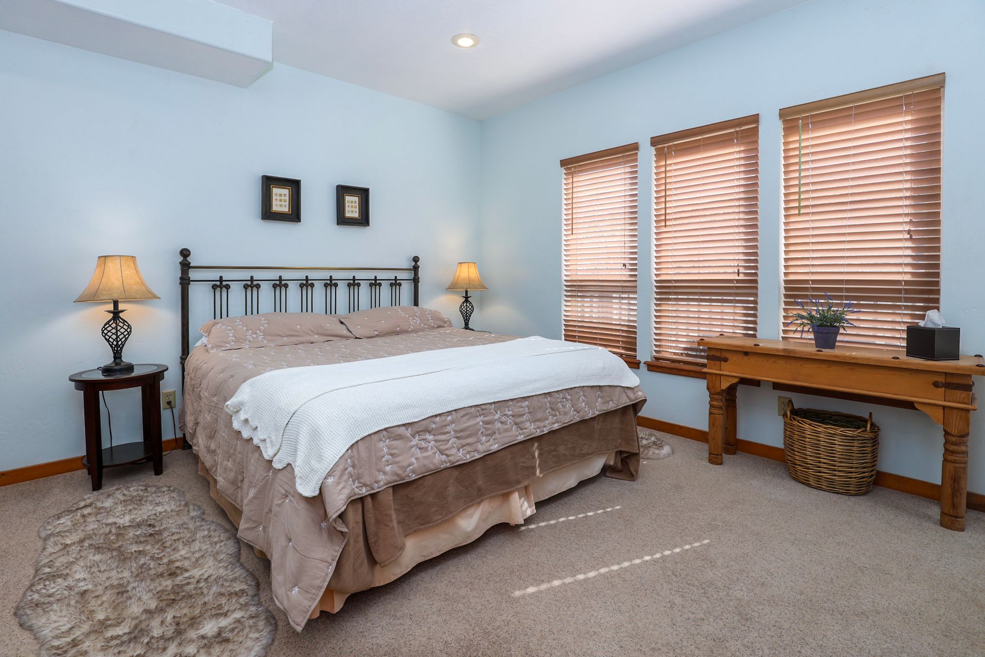 A bedroom with a king-sized bed, two side tables with lamps, a wooden console table, and three windows with wood blinds.