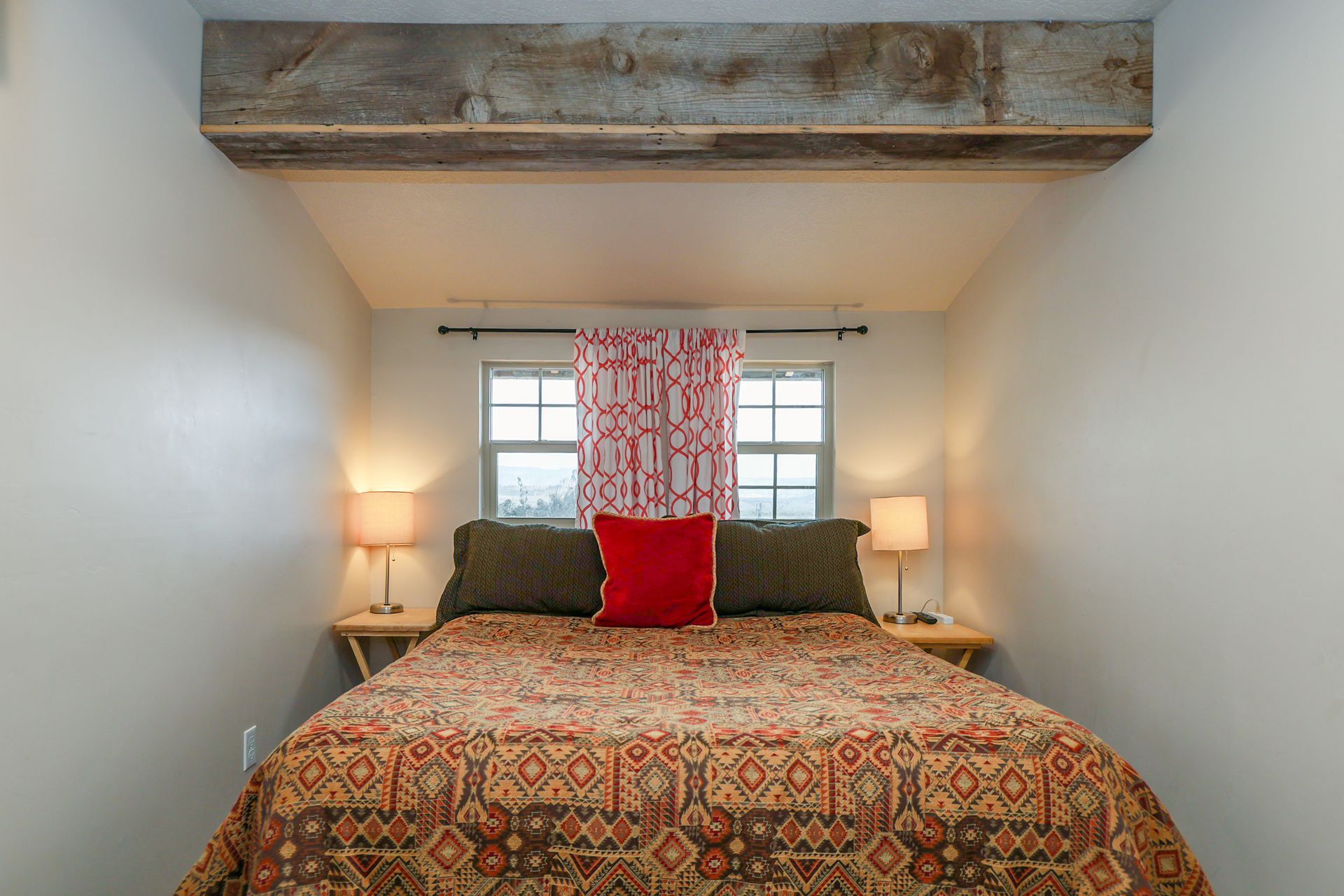A bedroom features a queen bed with a patterned quilt, dark pillows, and a red throw pillow below a window and wood beam.