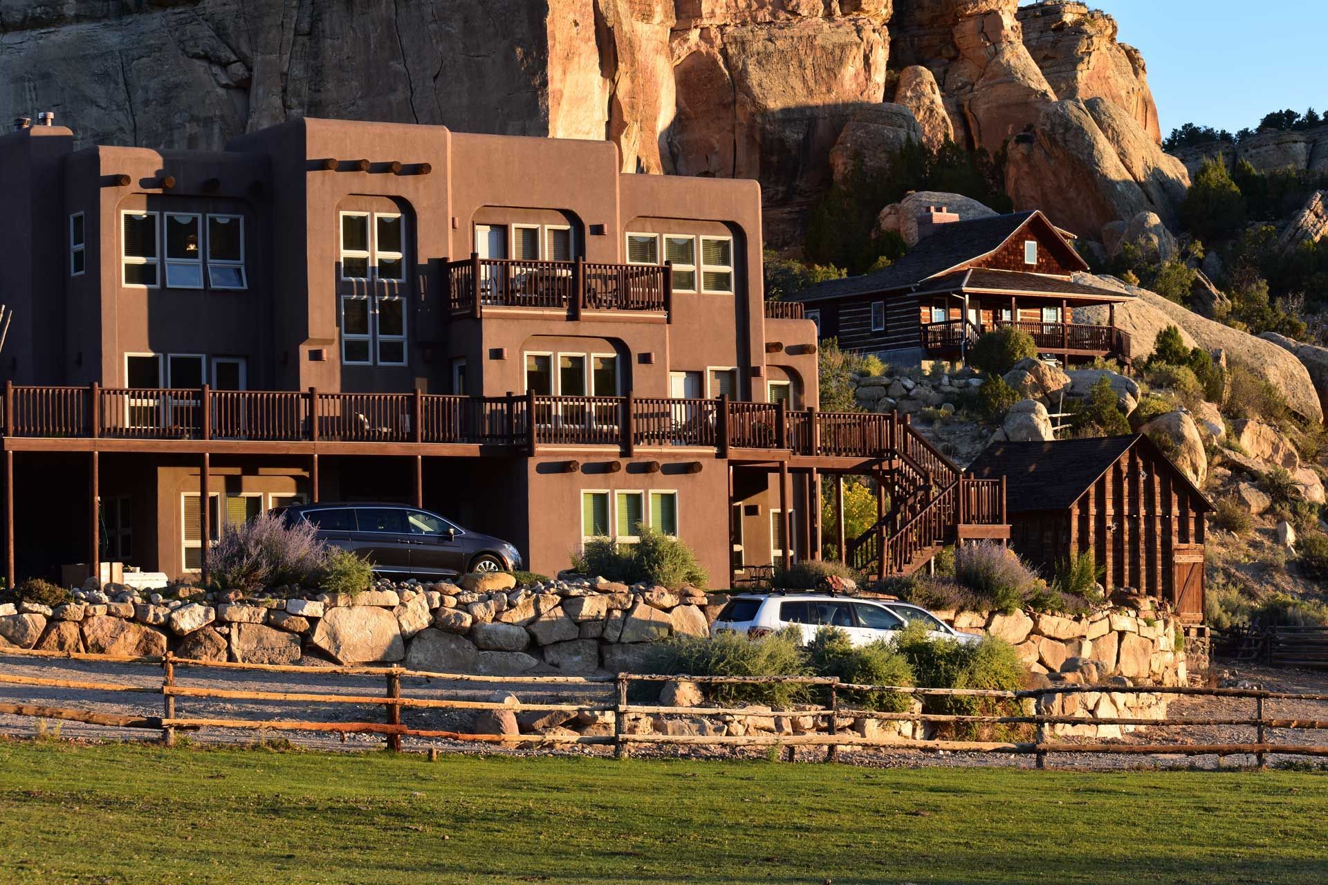 A large, multi-level brown stucco house with wooden decks sits at the base of a tall, rocky sandstone cliff at sunset.