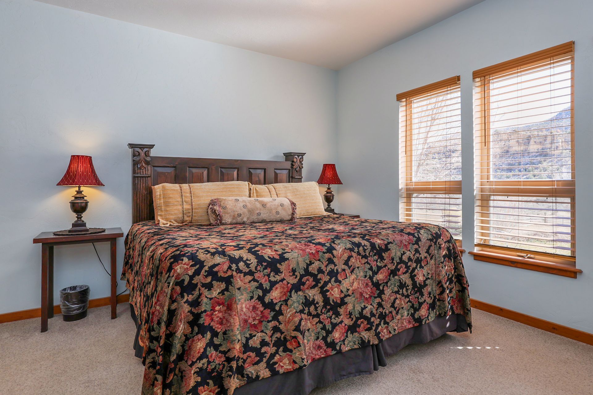 A king-sized bed with a floral patterned quilt sits against a light blue wall, with bedside lamps and two windows.