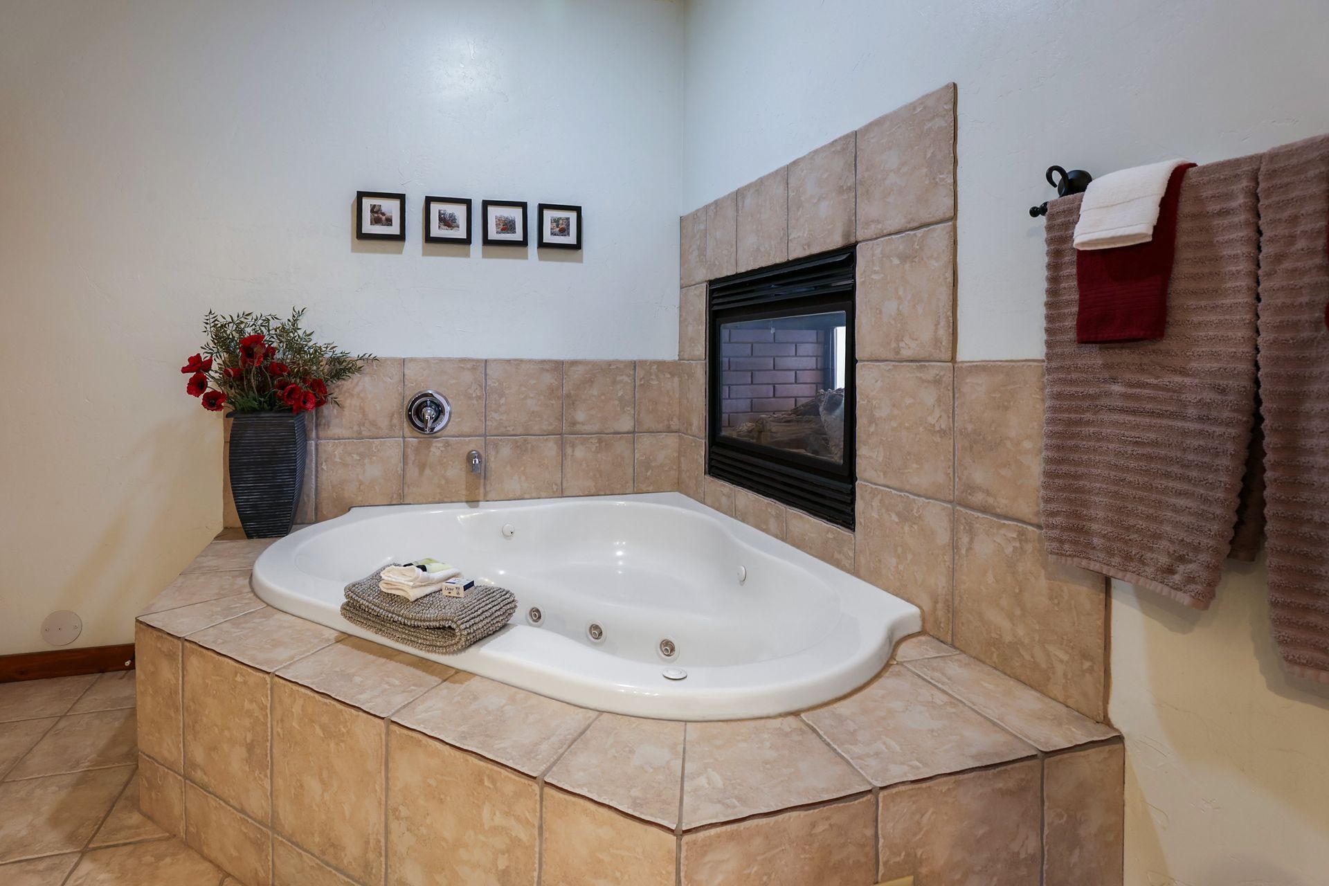 A corner jetted bathtub with beige tiling and a built-in fireplace, accented by a vase of red flowers and hanging towels.