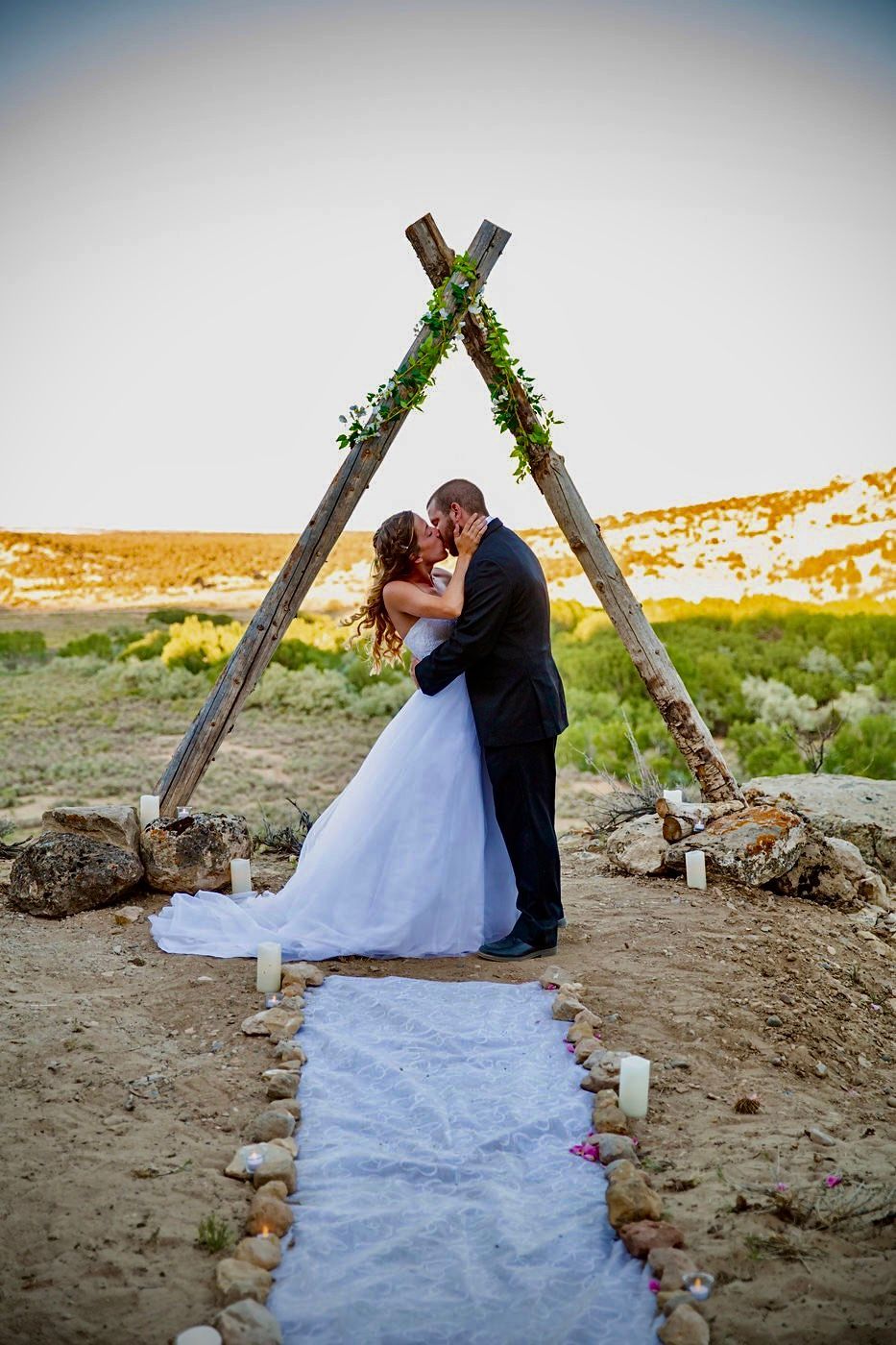 A couple kisses under a rustic, triangular wooden arch decorated with greenery on a dirt path with candles.