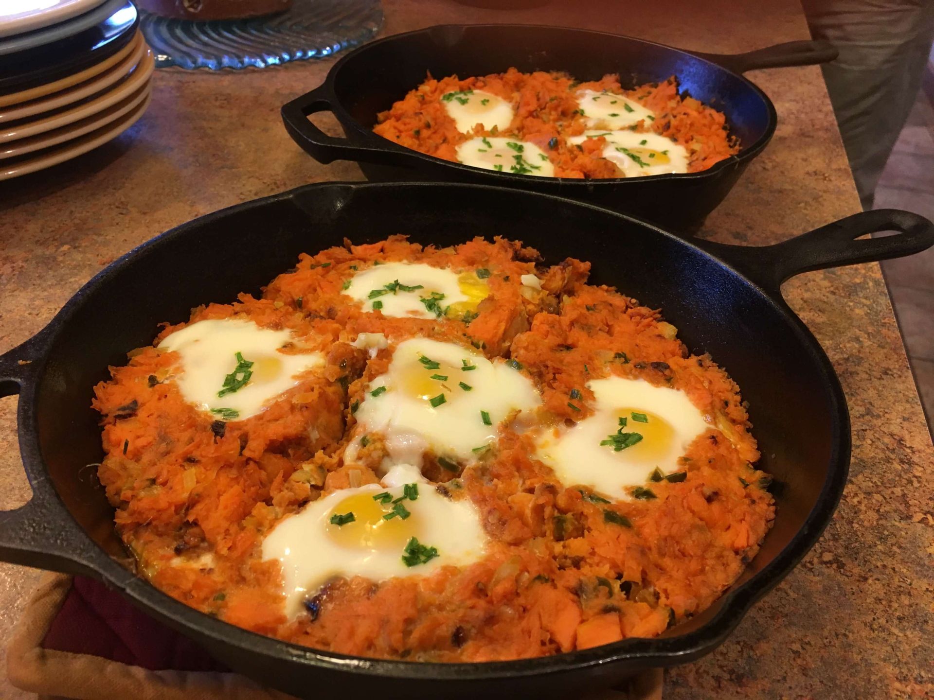 Two cast iron skillets containing sweet potato hash topped with sunny-side-up eggs and garnished with fresh herbs.