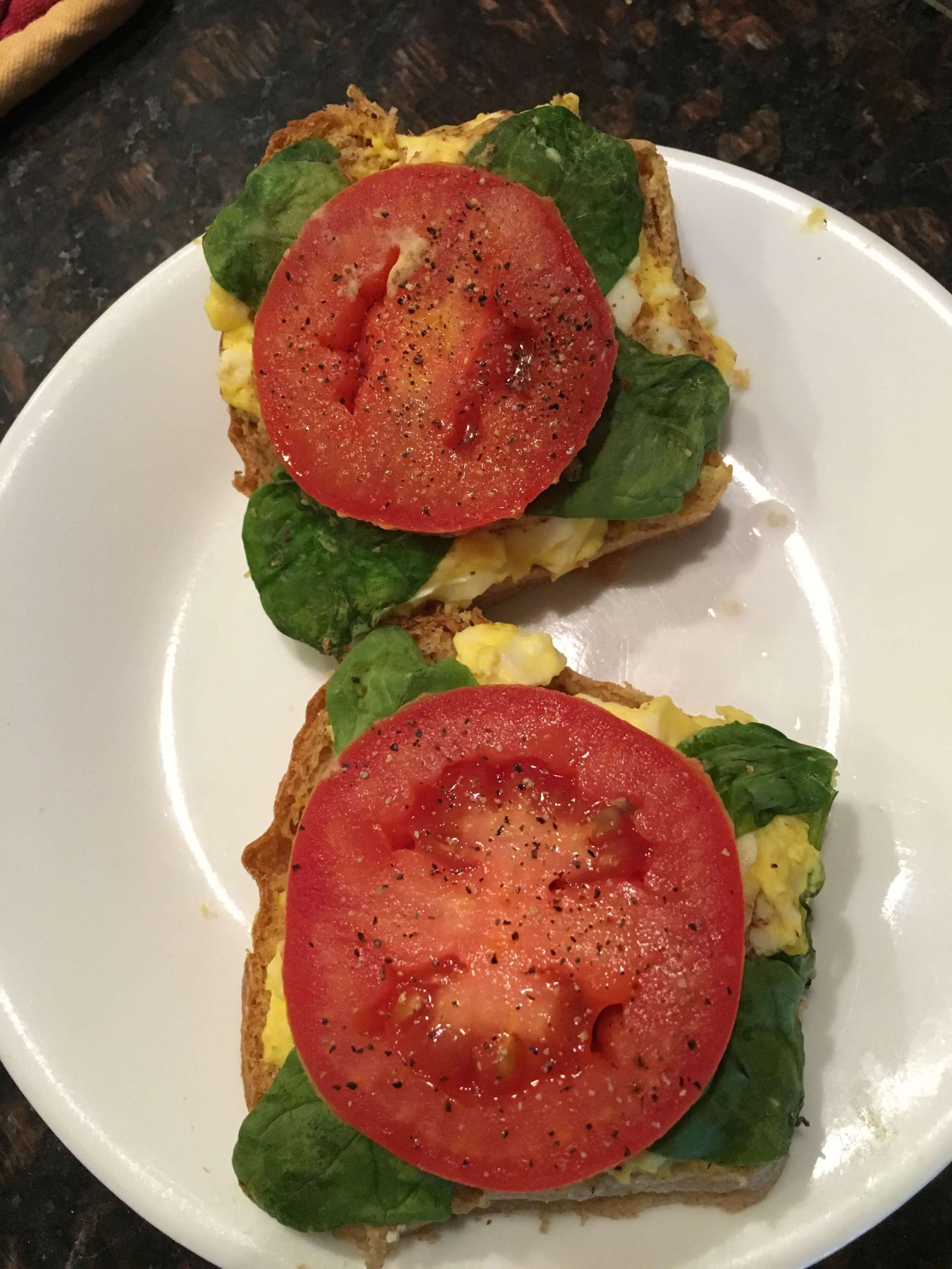 Two slices of toasted bread topped with scrambled eggs, fresh spinach, and thick-cut, peppered tomato slices on a plate.