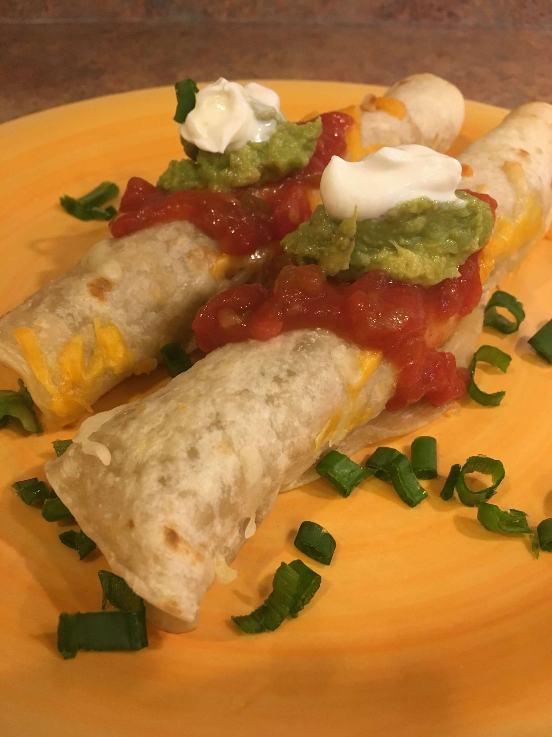 Two rolled tortillas topped with melted cheese, salsa, guacamole, sour cream, and green onions on a yellow plate.