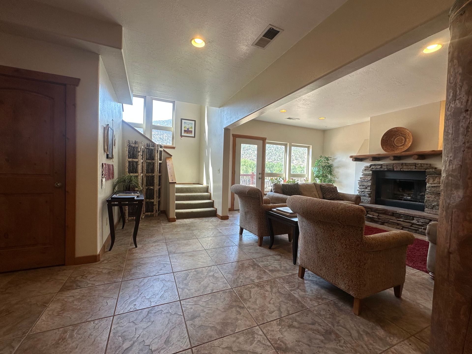 Spacious living area with tile floors, a stone fireplace, armchair seating, and stairs leading to a doorway.