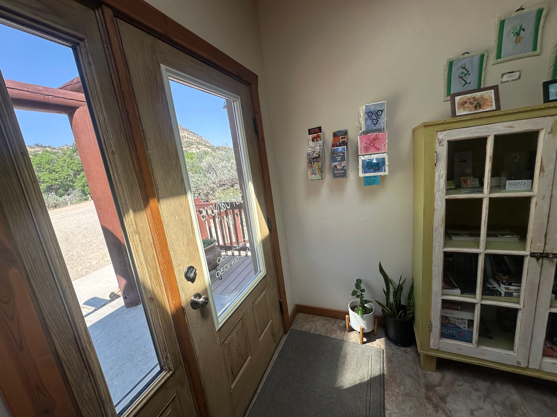 An entryway with double wooden doors leading outdoors, a beige wall with art, and a vintage yellow cabinet with glass.