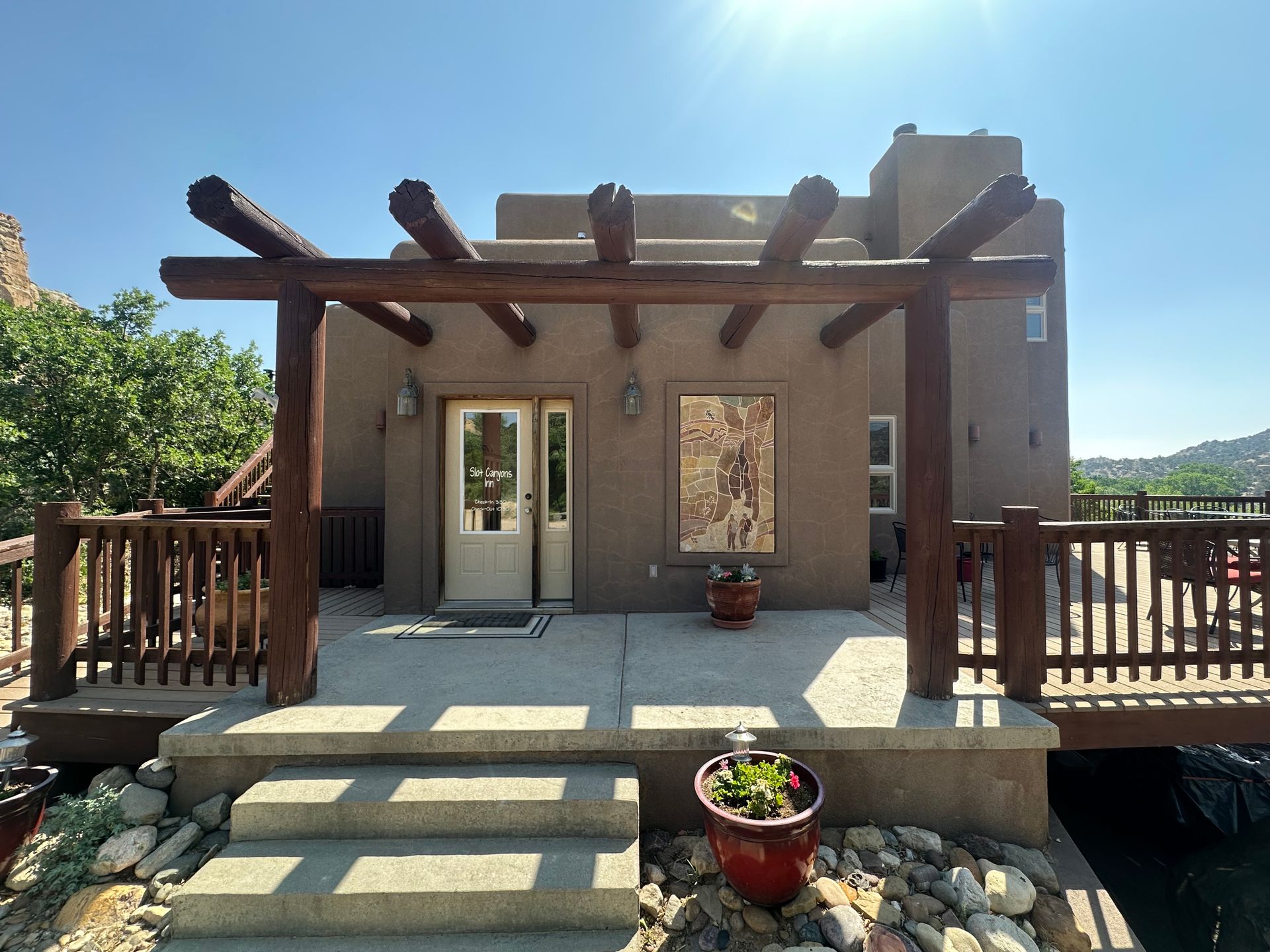 A brown stucco house with a wooden pergola over a concrete patio and steps, set against a bright, clear blue sky.