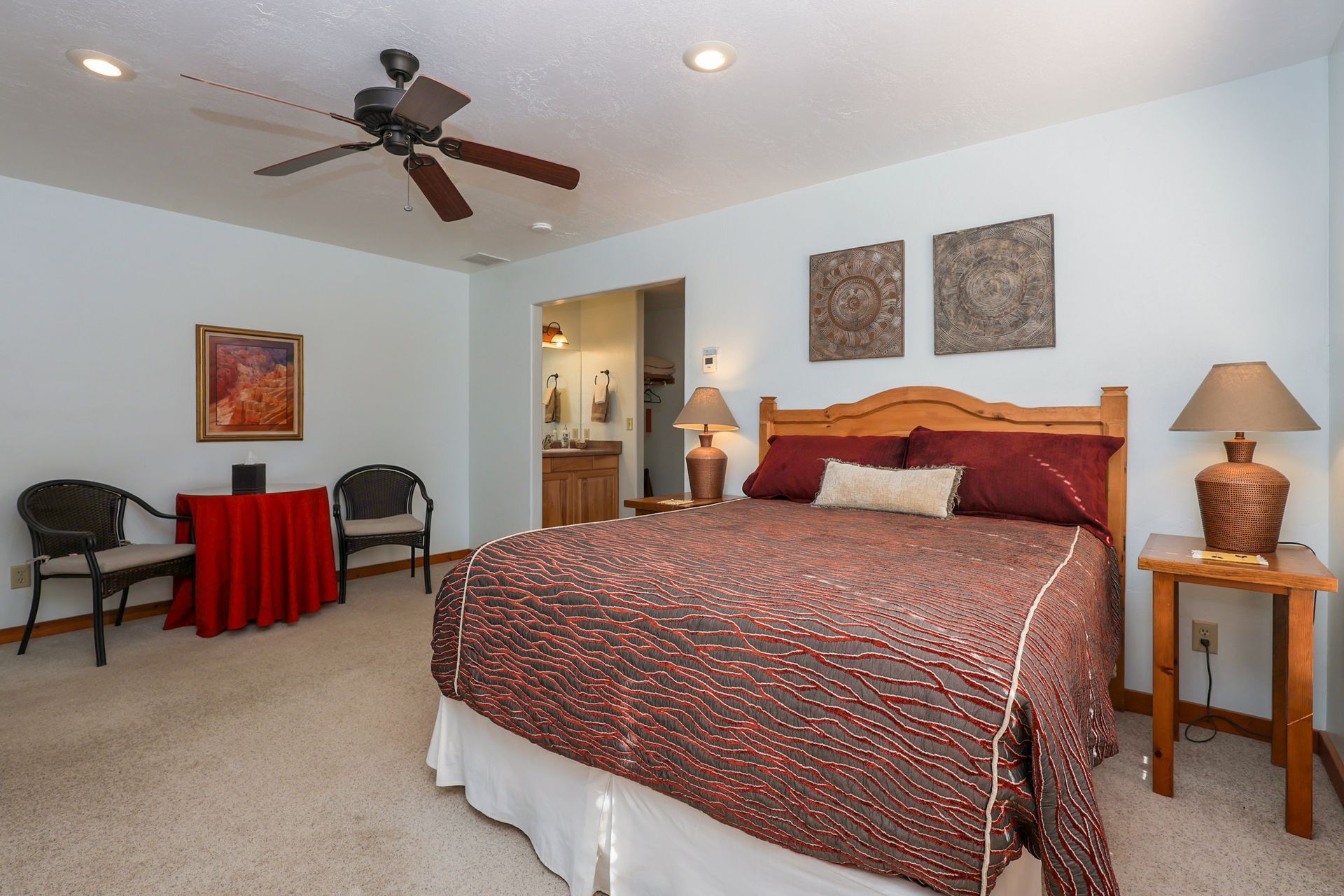 A bedroom with a queen bed featuring a patterned red spread, two side tables with lamps, and two chairs by a red table.