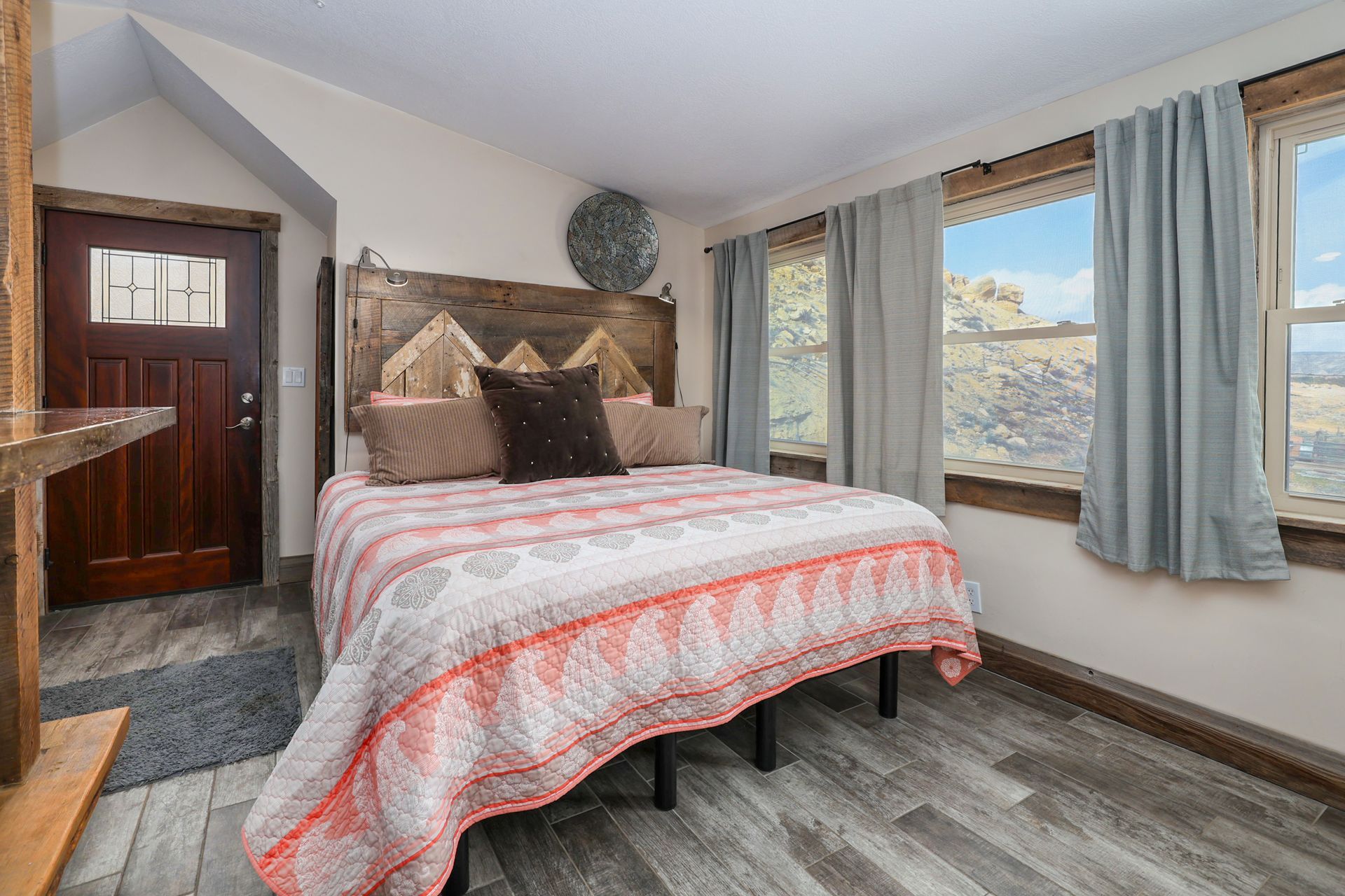 A rustic bedroom with a wooden headboard, patterned bedding, and large windows looking out onto a rocky landscape.