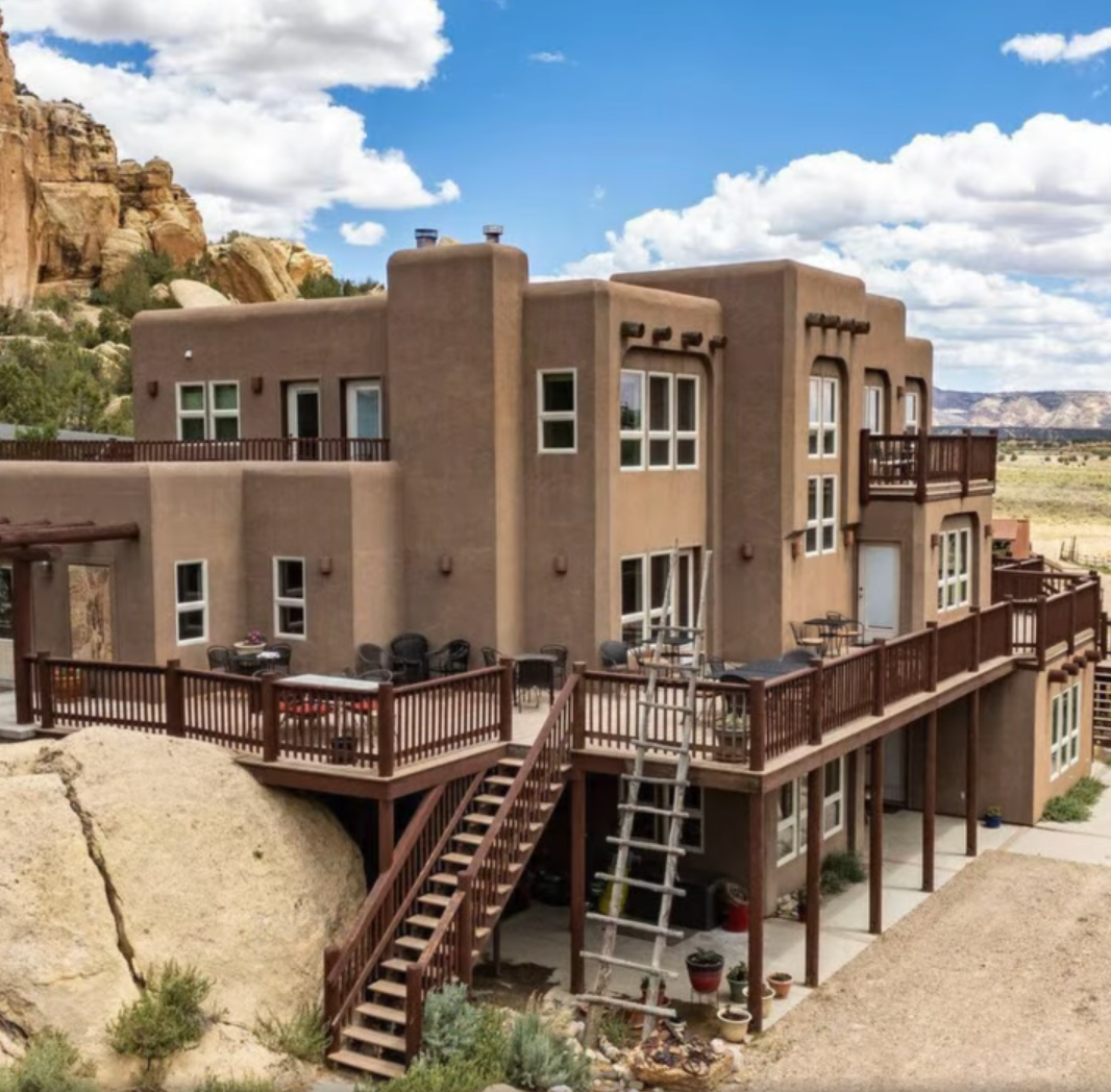 A multi-story, tan-colored stucco home with a desert-style exterior, large wooden deck, and balcony, set against rock.