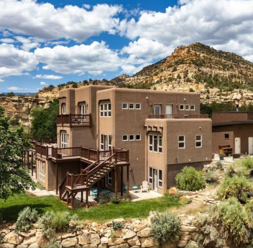 A three-story tan adobe house with wooden balconies, set against a rocky hillside under a blue sky with white clouds.