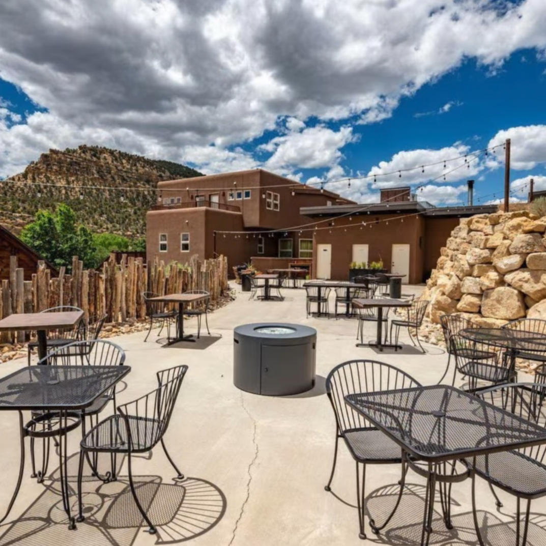 Outdoor patio with metal tables and chairs, a central fire pit, and a tan building against a rocky hillside under clouds.