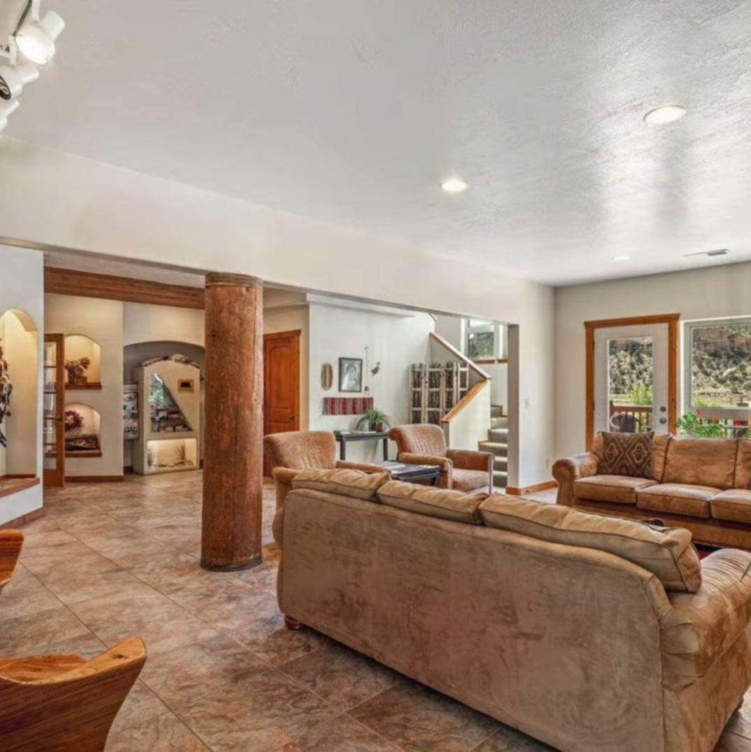 Spacious living room with tan sofas, tiled floors, a large wooden support pillar, and an open layout leading to stairs.