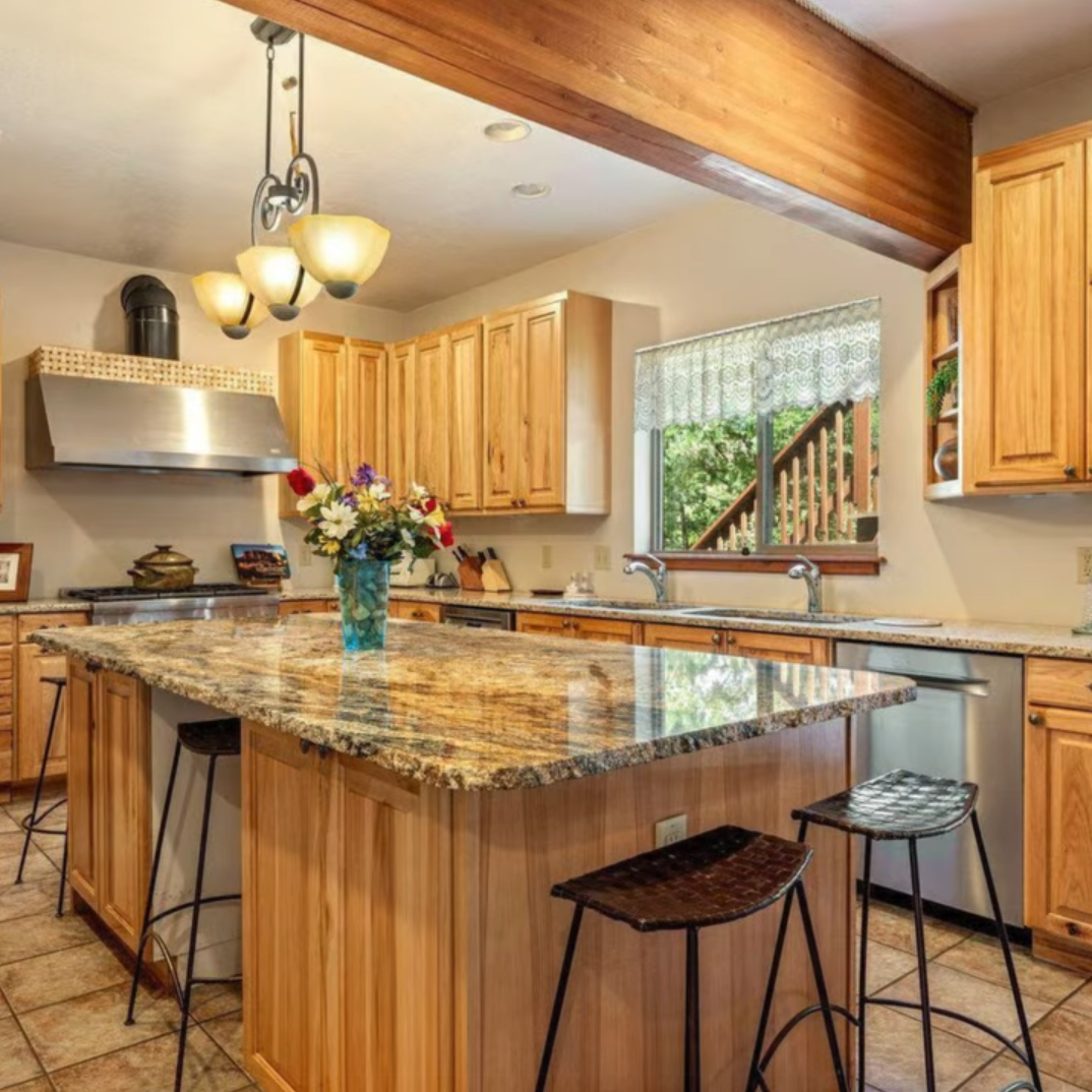 A kitchen with light wood cabinets, a granite-topped island with bar stools, stainless steel appliances, and a pendant light.
