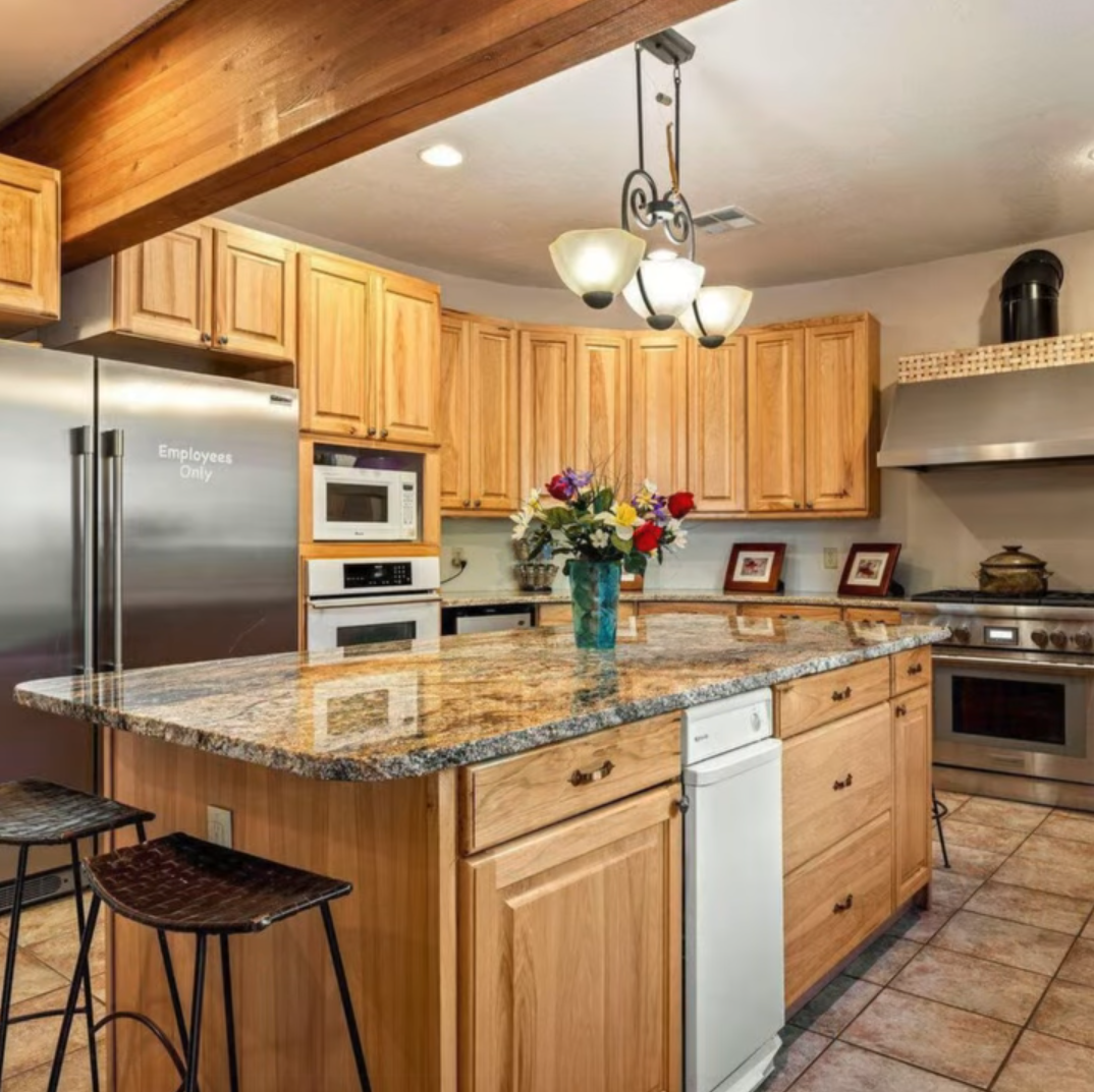A spacious, light-wood kitchen featuring a granite island, stainless steel appliances, and a hanging pendant chandelier.