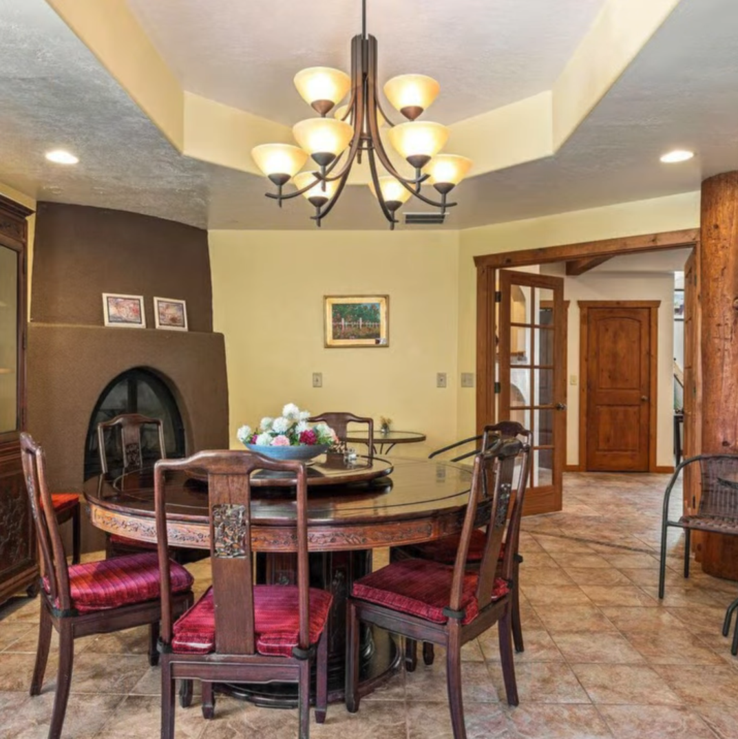 A formal dining room features a round wooden table with four chairs, a chandelier, a kiva-style fireplace, and tiled floor.