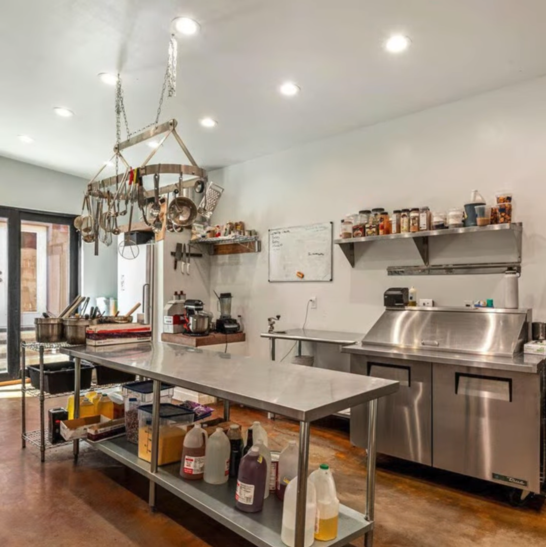 A professional kitchen with stainless steel prep tables, an overhead pot rack, shelving, and a large metal refrigerator.