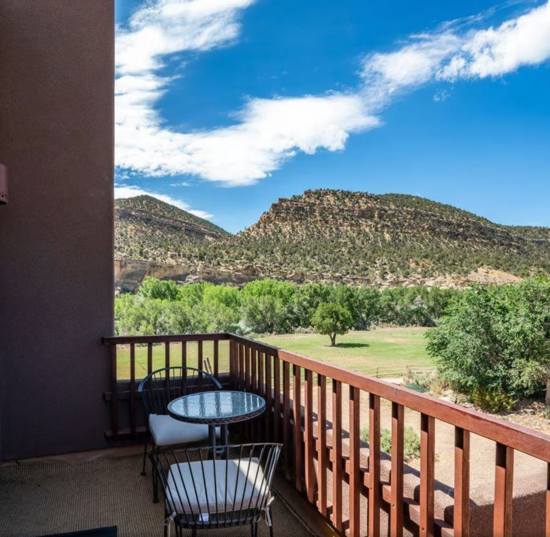 A balcony with a small round table and two chairs overlooking a green valley and rugged mountains under a blue sky.