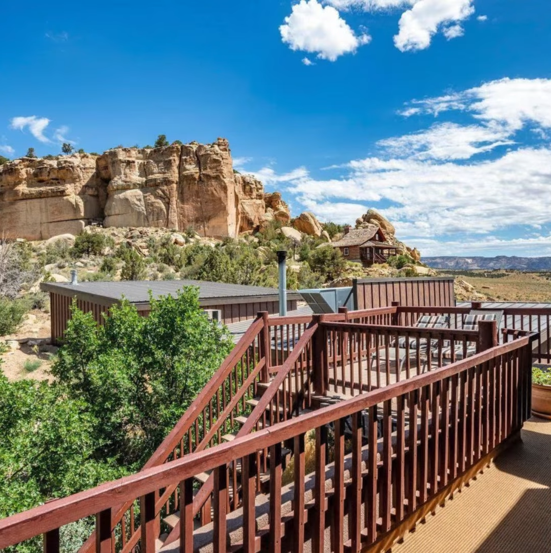 A wooden deck overlooks a desert landscape with sandstone cliffs under a blue sky with scattered clouds.