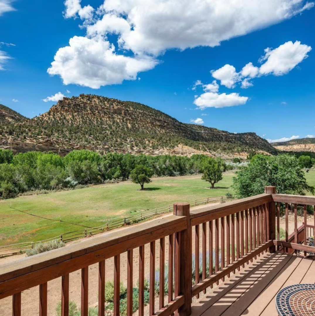 A scenic view from a wooden deck overlooking a lush green pasture, trees, and rocky mountains under a blue, cloudy sky.