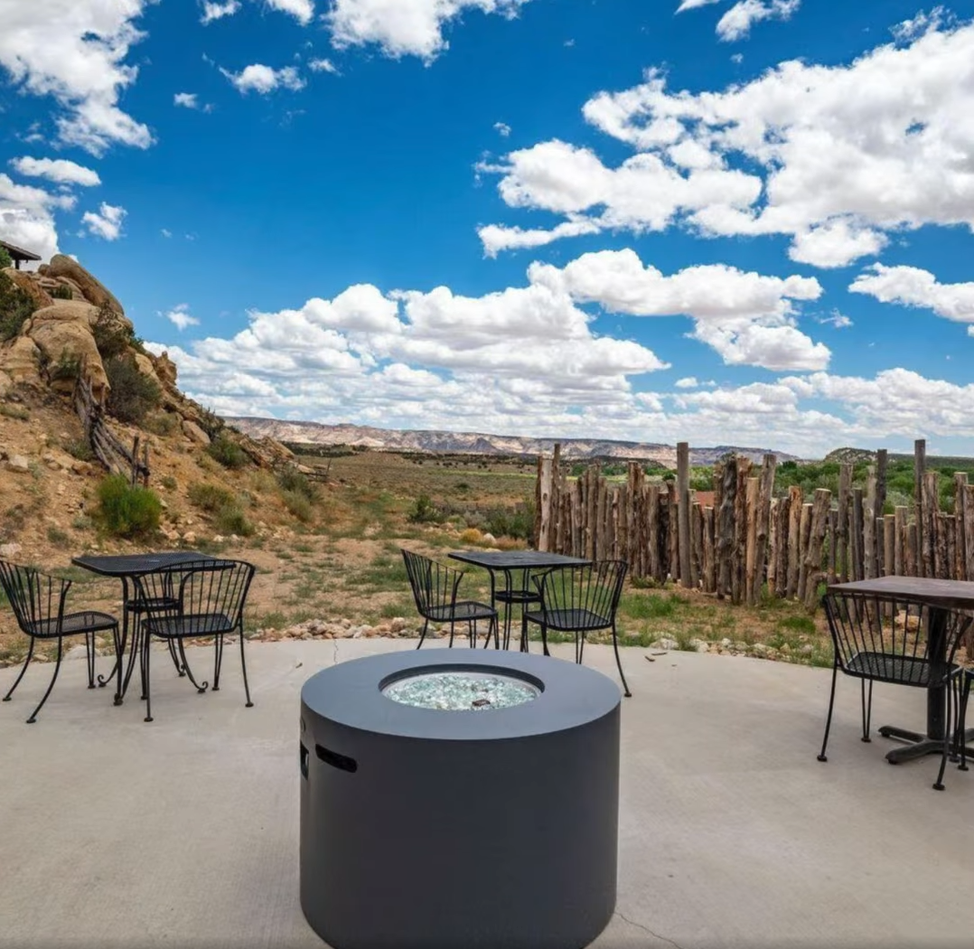 Outdoor patio with black metal tables and chairs, a cylindrical fire pit, and a rocky desert landscape under a blue sky.