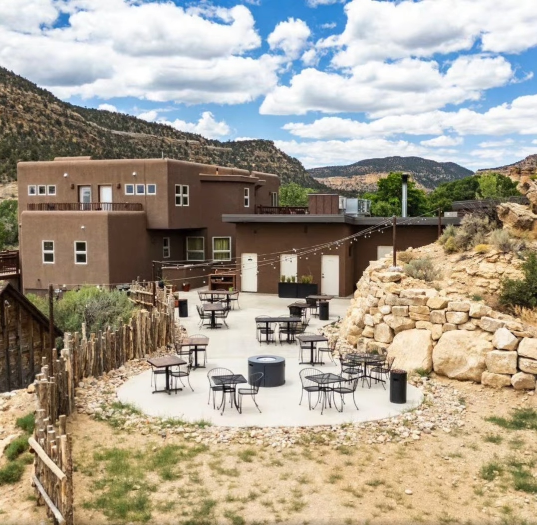 Outdoor patio with tables and chairs in front of a two-story brown stucco building against a scenic desert mountain.