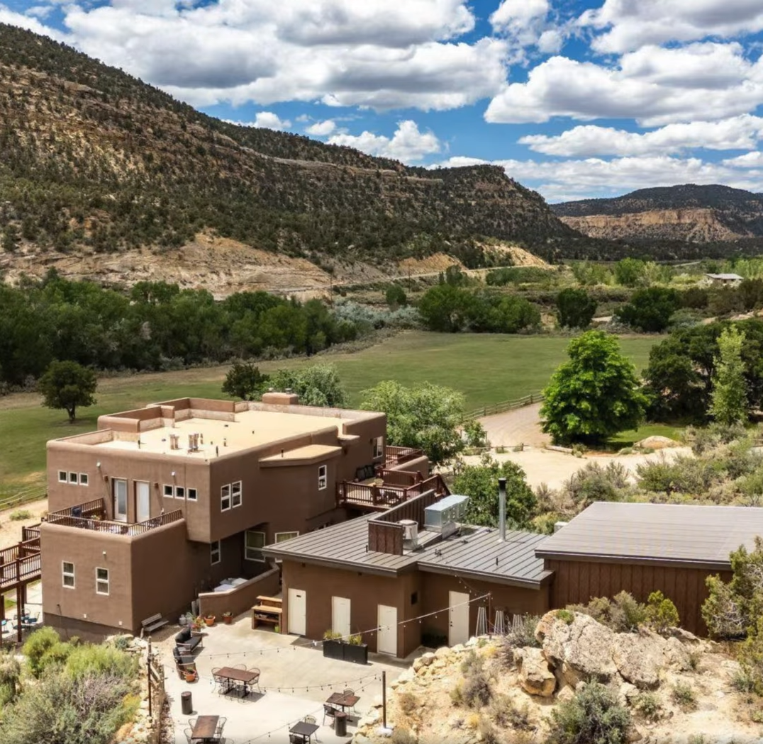 A multistory brown adobe-style building with a flat roof sits in a desert landscape against large hills under a blue sky.