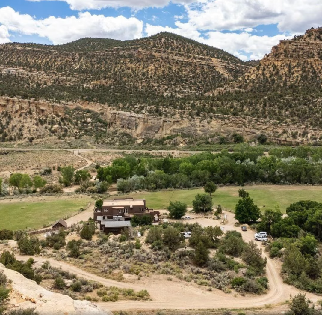 A brown building sits in a green valley before tall, rugged, tan-colored mountains under a bright, cloudy sky.