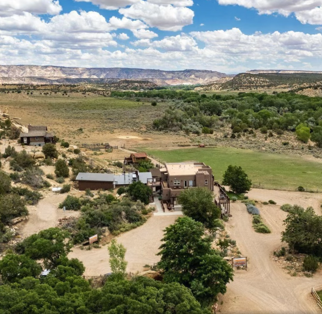 An aerial view of a tan, multi-level house nestled in a desert landscape with scrub brush, green trees, and rocky mesas.