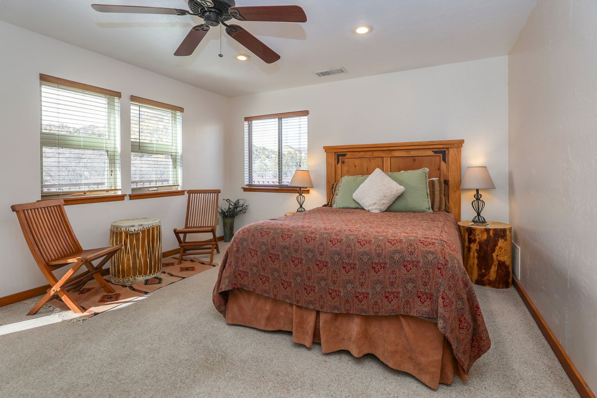 A bedroom with a queen bed, two wooden chairs by a window, two stump-style side tables, and a ceiling fan.