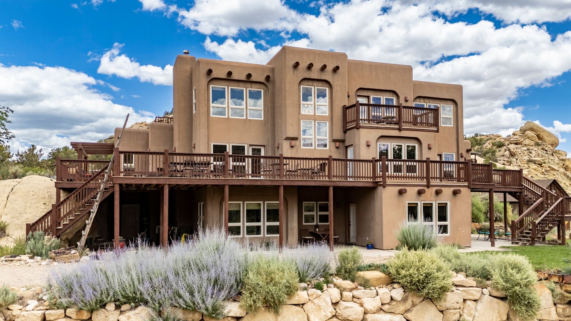 A three-story, tan-colored southwestern-style house with a wooden deck, set against a rocky, desert-like landscape.