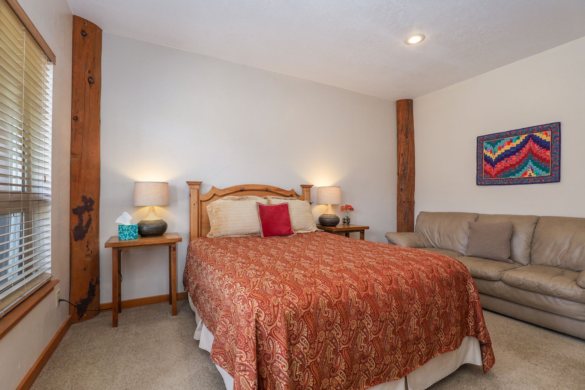 A hotel-style bedroom features a bed with an orange patterned quilt, two lamps on nightstands, and a light-colored sofa.