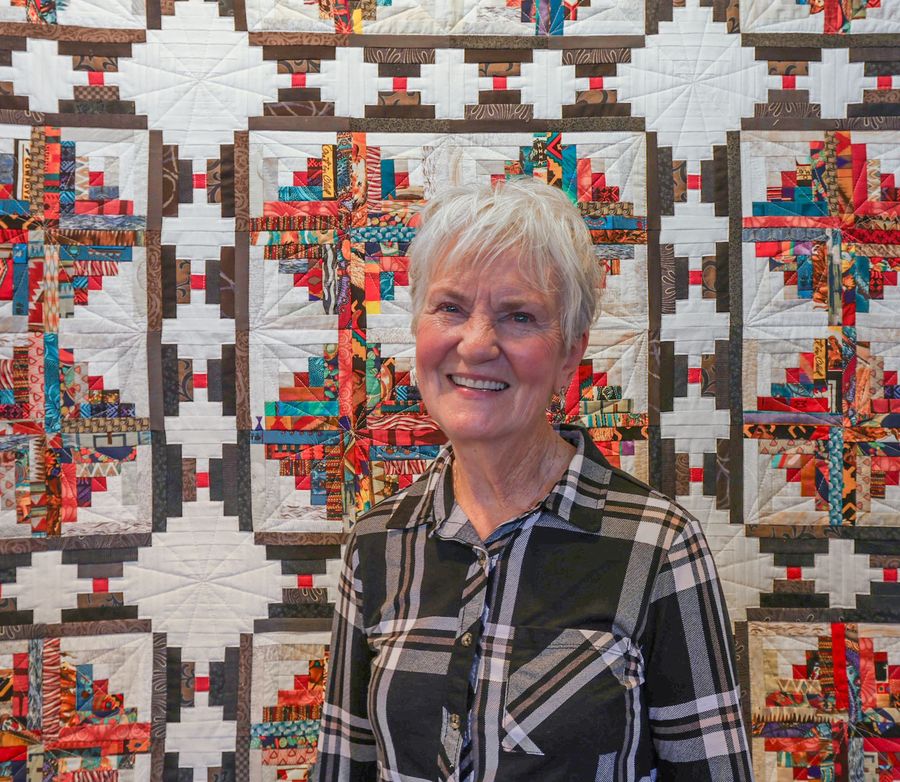A smiling person in a black and white plaid shirt stands in front of a colorful, patterned quilt wall hanging.