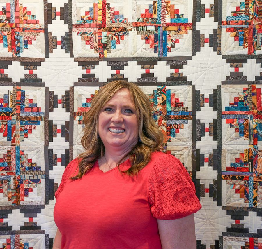 A smiling person in a red top stands in front of a colorful, patterned patchwork quilt.