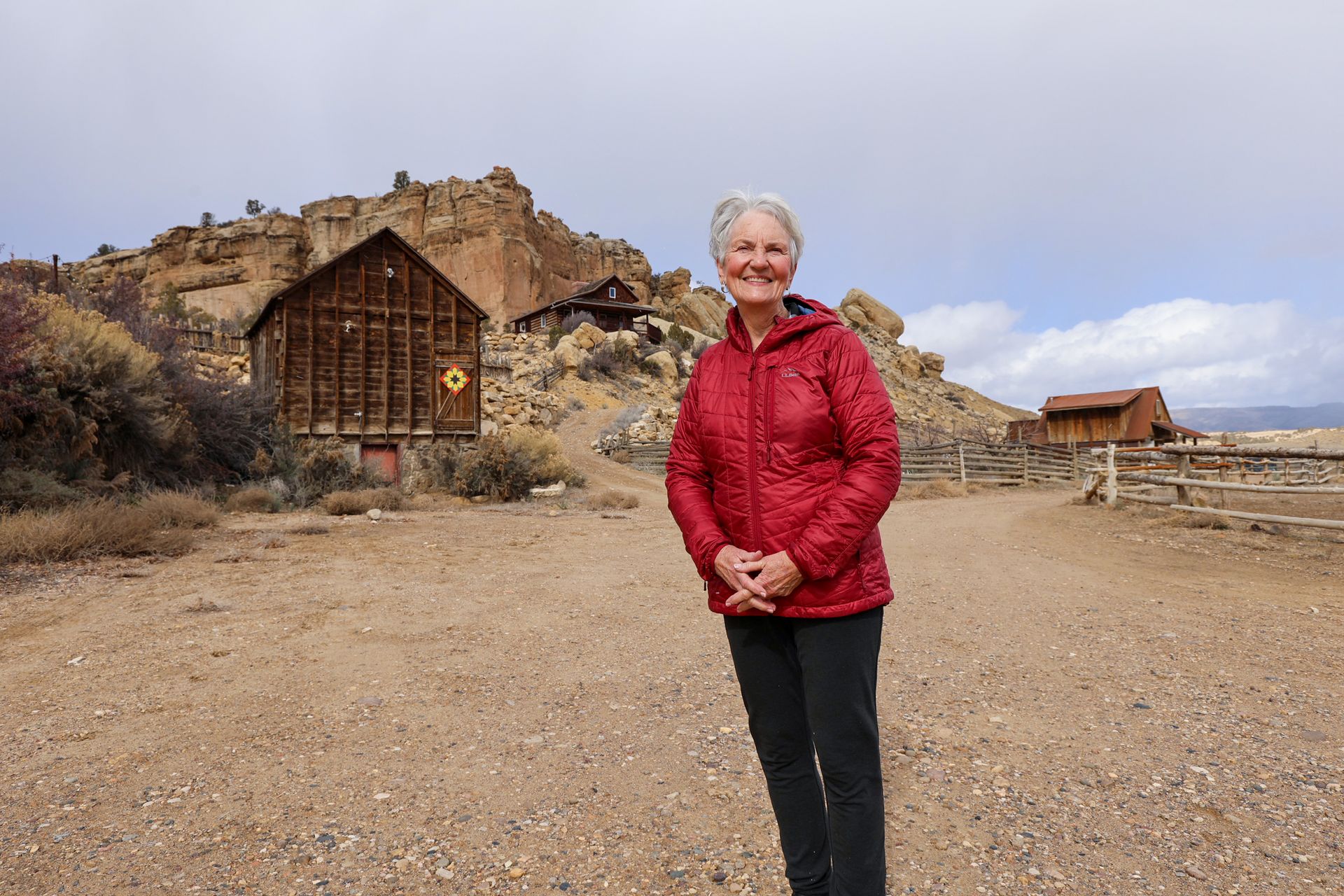 A person in a red puffer jacket standing on a dirt path in front of a rustic wooden building and rocky hillside.