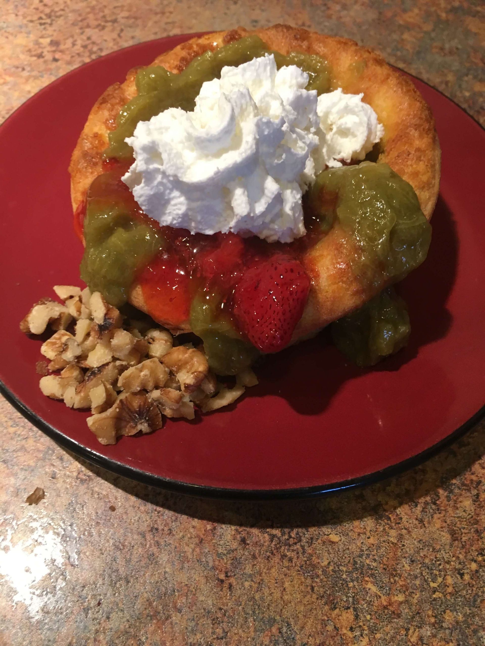 A round fried pastry on a red plate, topped with strawberry jam, green fruit compote, whipped cream, and chopped walnuts.