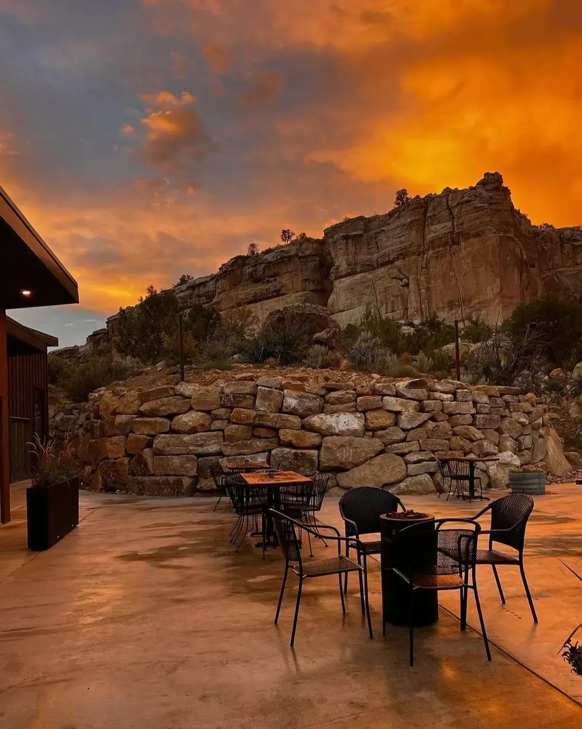 Patio seating area at sunset with metal tables and chairs before a large, rocky cliff face under a vibrant orange sky.