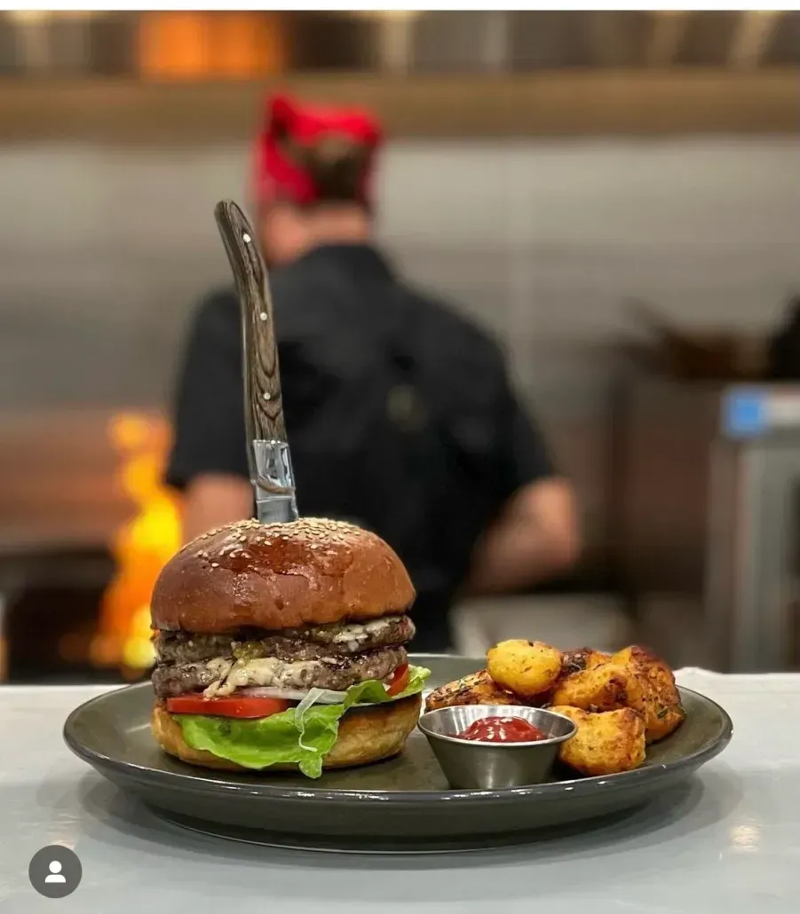 A double cheeseburger with a knife stuck in the top bun, served with tater tots and ketchup, with a chef in the background.