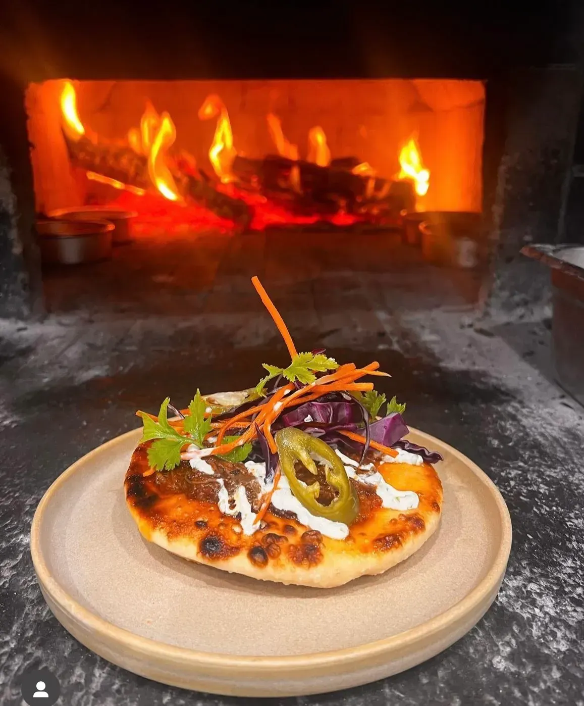 A garnished flatbread topped with jalapeño, shredded vegetables, and crema, set on a plate in front of a wood-fired oven.
