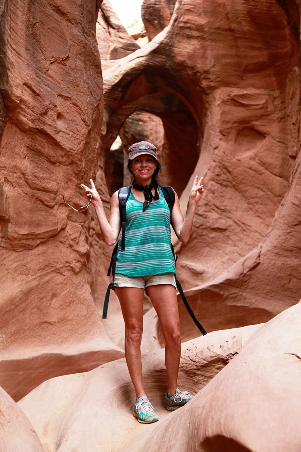 A person in a hat and tank top stands between narrow, curving orange sandstone walls, smiling and making peace signs.